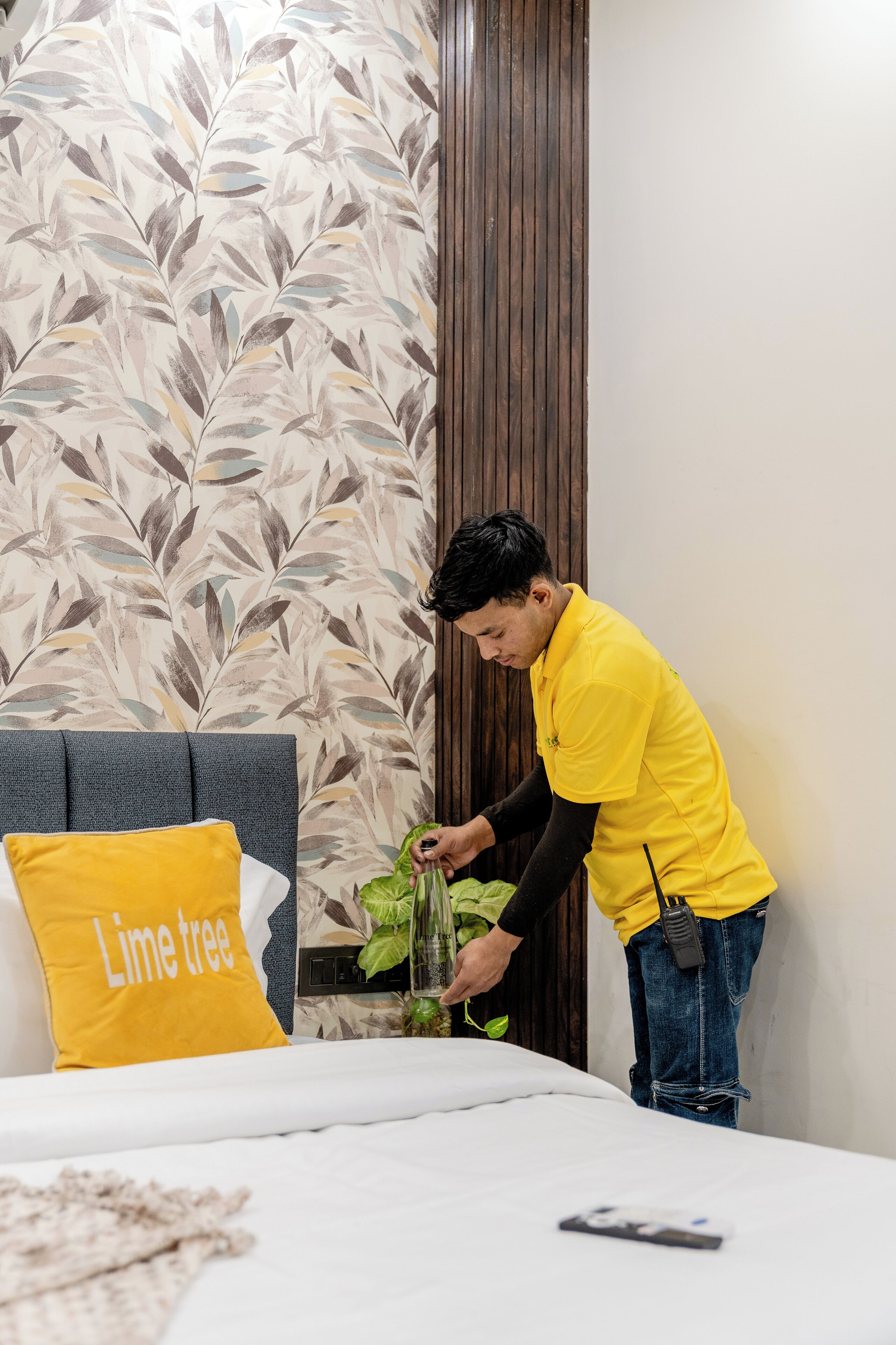 A hotel room with a bed, a pillow labeled "Lime tree," a bedside plant, and a wooden panel wall.