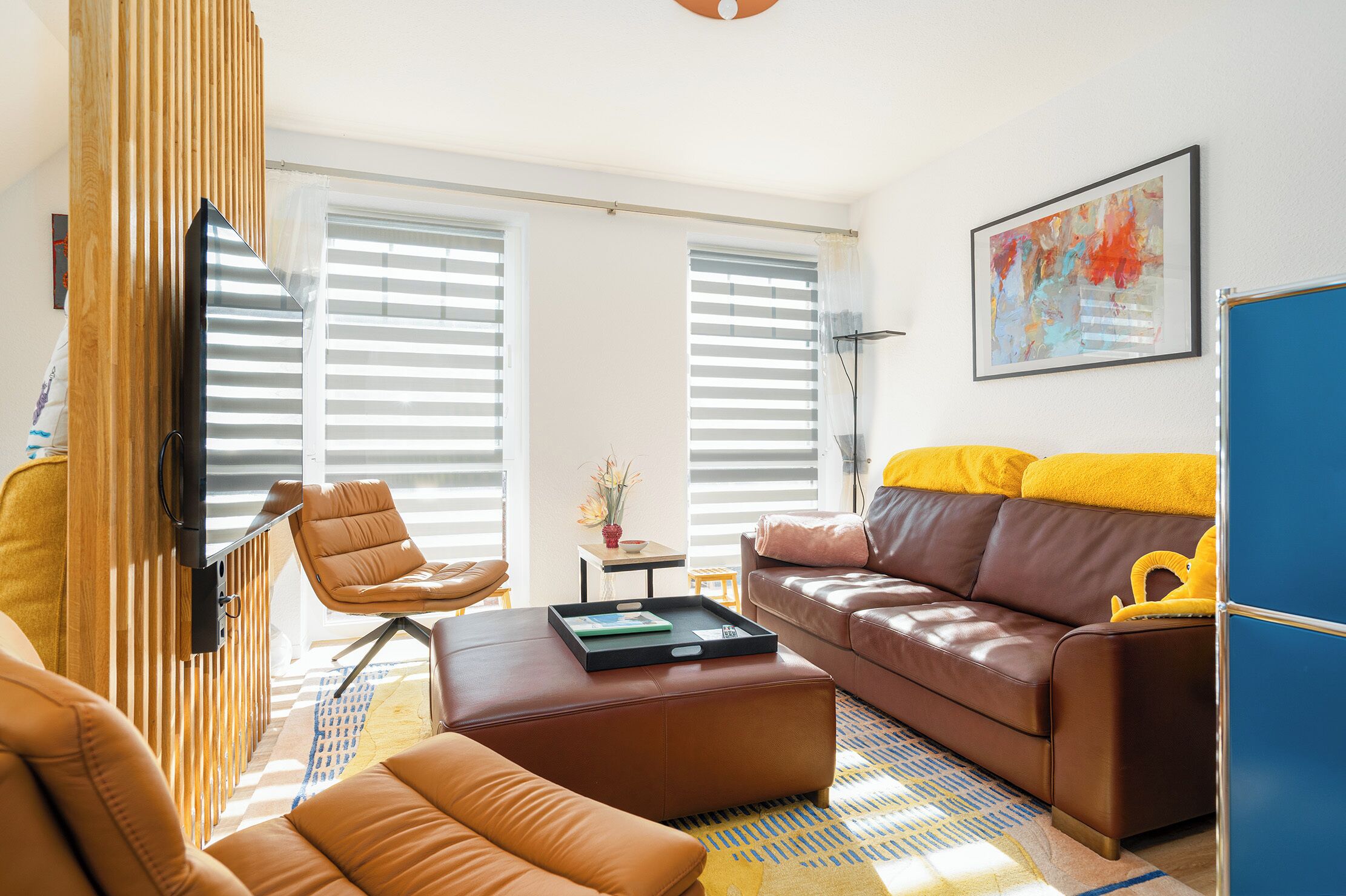 Un salon moderne avec un canapé en cuir marron, un fauteuil en cuir marron assorti et un pouf assorti. Un téléviseur à écran plat est fixé sur un panneau en bois, il y a une petite table avec un vase, et un tableau abstrait aux couleurs vives est accroché au mur.
