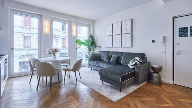 A modern living room with a grey sofa, a round coffee table, and two chairs. There is a large window with a view of buildings outside.
