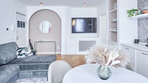 A modern living room with a grey sofa, a round coffee table with a vase of dried pampas grass, a flat-screen TV mounted on a white wall, and a kitchen area with white cabinets and shelves.