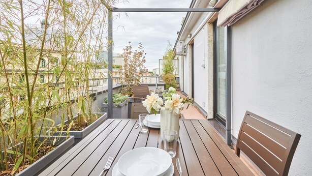 Un balcon en bois avec une table dressée pour deux personnes, un banc et un vase rempli de fleurs.