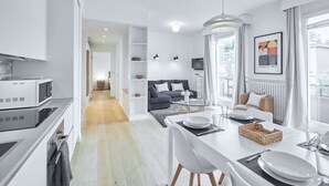 Une cuisine moderne avec des armoires blanches, un four à micro-ondes et une cuisinière. On y trouve un coin repas avec une table blanche, des chaises et un buffet. Un salon avec un canapé et un téléviseur est visible au fond.