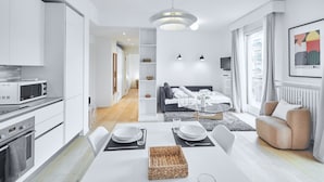 Une cuisine moderne avec des meubles blancs, une table à manger pour quatre personnes et un coin salon avec un canapé et un téléviseur.