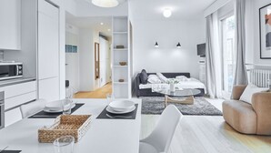 Un salon moderne et bien éclairé, avec un canapé, un coin repas avec une table dressée pour deux personnes, et un coin cuisine équipé d’un four à micro-ondes.