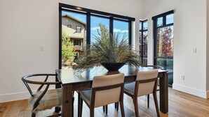 A dining area with a wooden table and chairs, a potted plant, and large windows offering a view of a residential building and trees. - Townhome w/Hot Tub | Walk to Ski Lift (Park City)