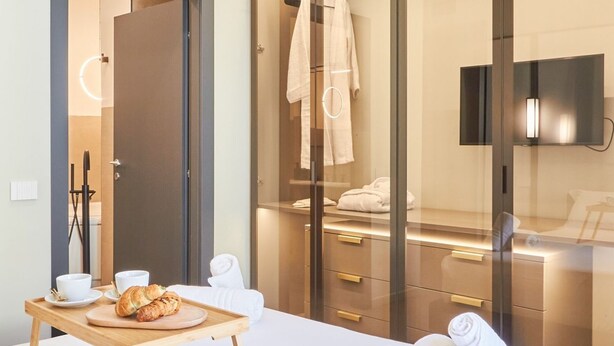 A modern hotel room with a wooden bedside table, a bed with white linens, a glass-enclosed walk-in closet, and a mounted TV.