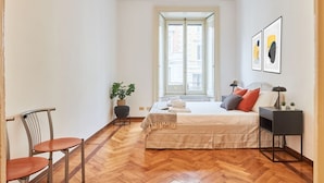 Une chambre avec un parquet en bois disposé en chevrons, un lit avec une literie blanche et de nombreux oreillers, une table de chevet avec une lampe, une chaise et une fenêtre donnant sur des immeubles.