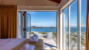 A bedroom with a large bed, two towels, and a view of a balcony with a wooden deck and a scenic lake.