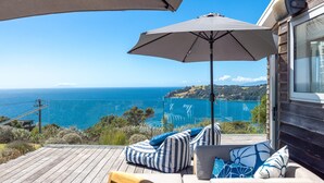 A wooden deck with outdoor seating, a large umbrella, and a view of the ocean and hills.