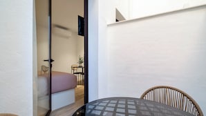Ett modernt rum med en glasdörr som leder in till ett sovrum, ett runt glasbord, flätade stolar och en liten krukväxt.
