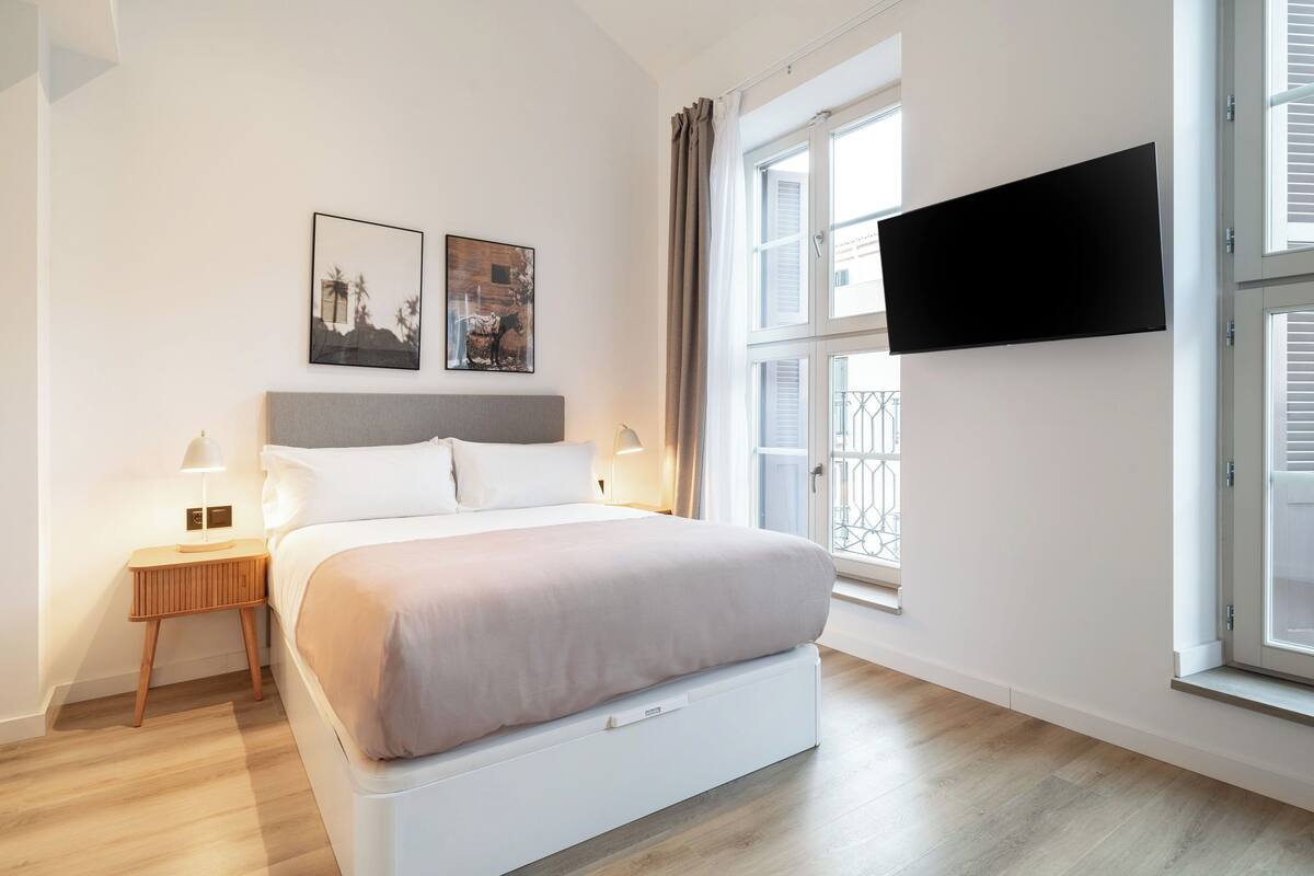 Ein modernes Schlafzimmer mit Bett, Nachttischen, einem an der Wand montierten Fernseher und großen Fenstern.