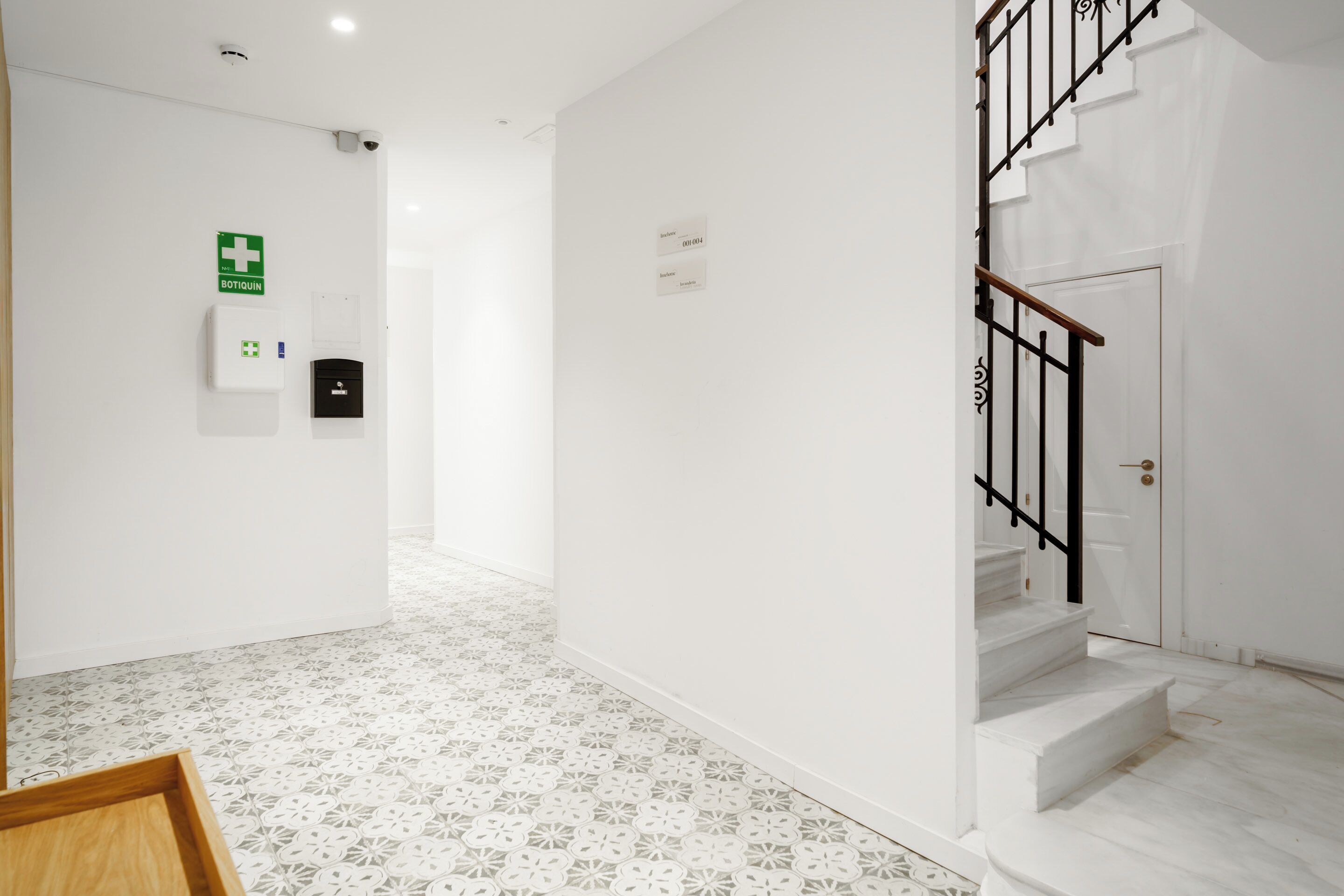 A clean, modern hallway with a white floor and walls, a staircase with black railings, and a visible emergency exit sign.