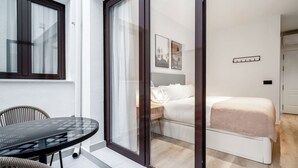 Ett modernt rum med en glasdörr som leder ut till en balkong, en säng med en vit sänggavel och ett litet bord med stolar.