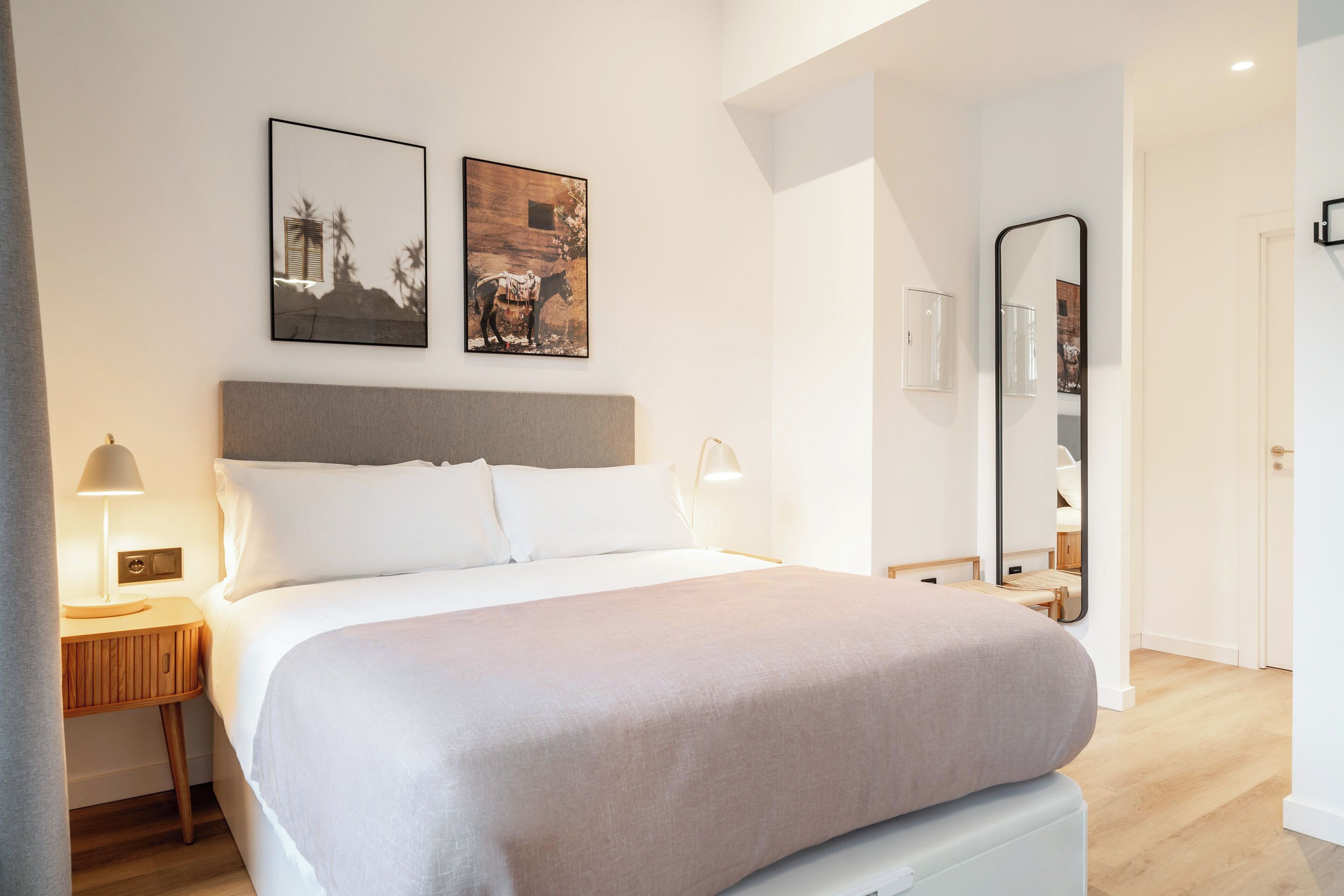 Quarto de hotel moderno com uma cama grande, uma mesa de cabeceira com abajur e duas imagens emolduradas na parede.