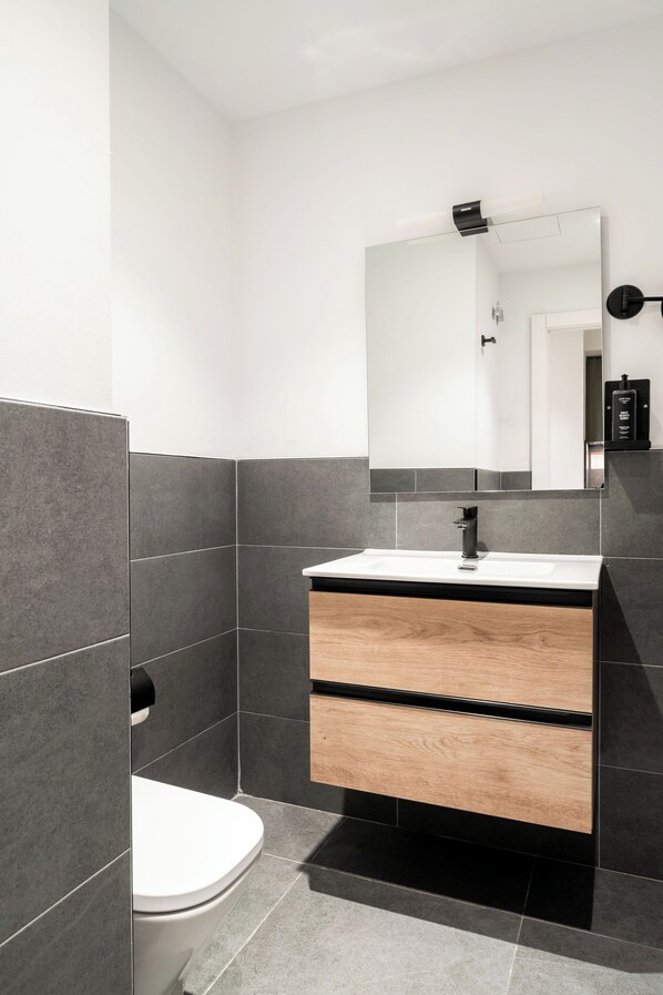 Un baño moderno con lavamanos blanco, baldosas oscuras y un mueble de tocador de madera.
