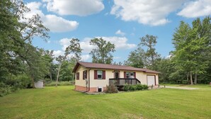 A single-story house with a front porch, a garage, and a small shed in a grassy yard surrounded by trees.