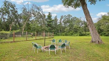 Un espace extérieur verdoyant, avec des chaises disposées en cercle autour d’un foyer, le tout entouré d’arbres et d’une clôture.