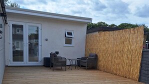 A covered patio with a wooden deck, two wicker chairs, a small table, and a glass door leading to a building. - Lodge - Sleeps 2 - Pet Friendly - Parking - Patio (Sidcup)