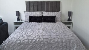 A neatly made bed with a grey headboard, two bedside tables with lamps, and a striped ottoman. - Lodge - Sleeps 2 - Pet Friendly - Parking - Patio (Sidcup)