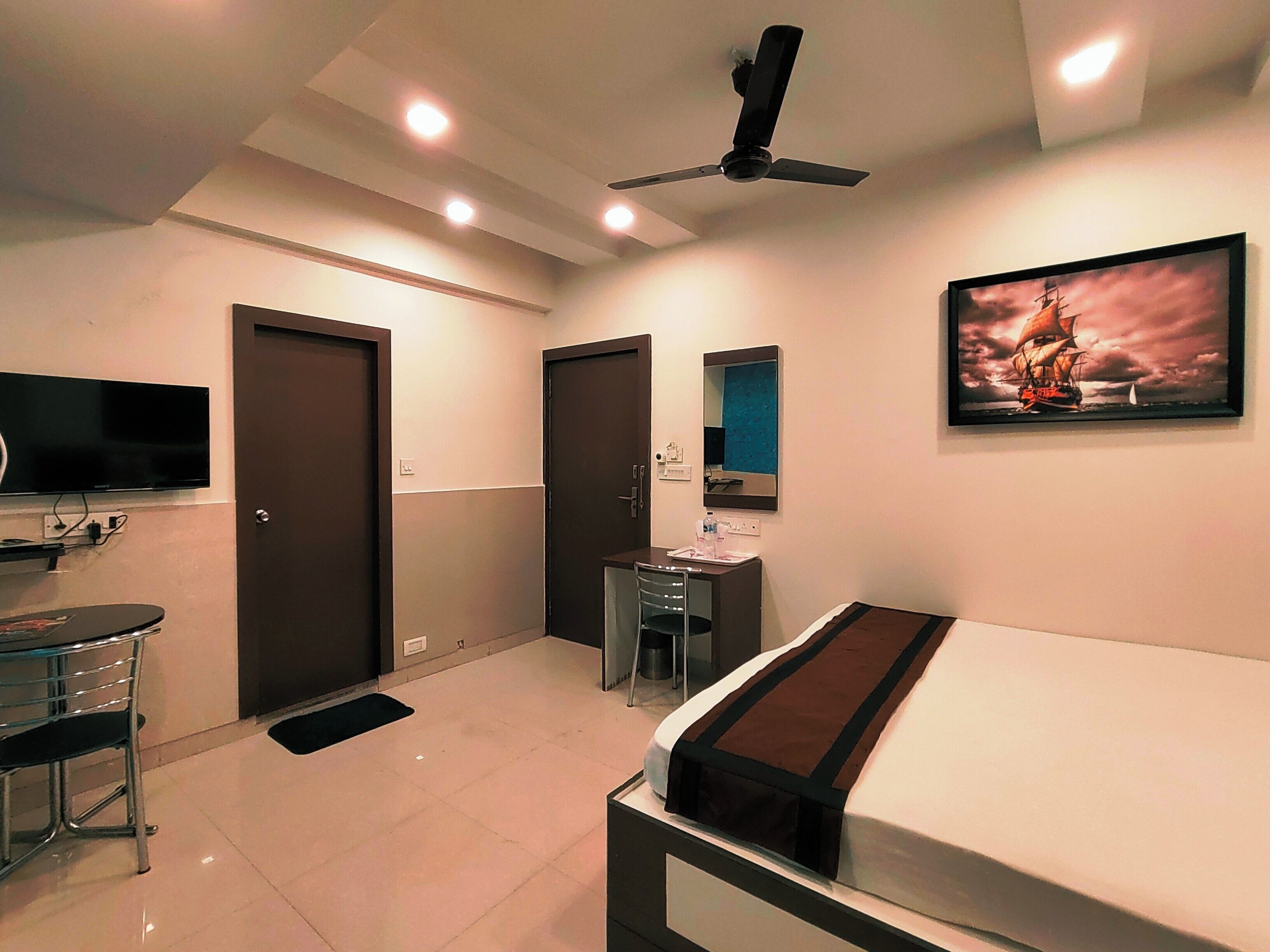 A hotel room with a bed, a desk, a chair, a TV, a ceiling fan, and a painting on the wall.