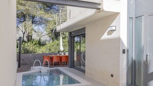 Un área de piscina moderna al aire libre con una mesa y sillas, rodeada por un muro de piedra y árboles.