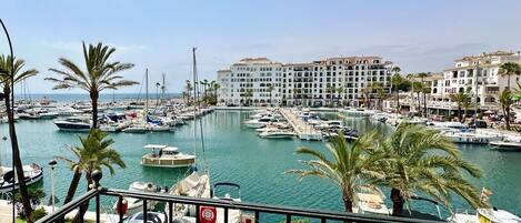 A marina with boats docked, palm trees, and a building with multiple balconies in the background.