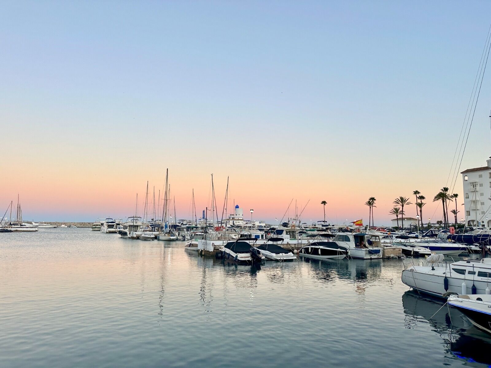 A marina with multiple boats docked, a calm water surface, and a clear sky.