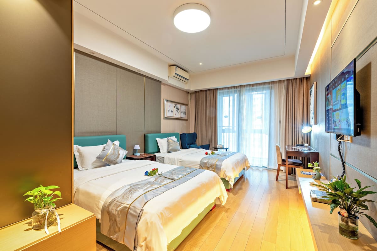 A hotel room with two beds, a desk, a TV, and a large window with curtains.