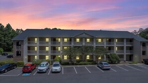 Ein mehrstöckiges Motel mit Parkplatz, das eine Reihe von einstöckigen Gebäuden mit Balkonen, einem zentralen Innenhof und einem Sonnenuntergangshimmel zeigt.
