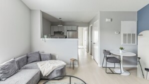 En moderne stue med en grå sofa, et lille rundt bord og en hvid stol. Et køkken er synligt i baggrunden med hvide skabe og rustfrie stålgrej.