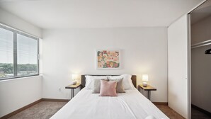 Ein Schlafzimmer mit einem Bett, zwei Nachttischen mit Lampen, einem Fenster mit Jalousien, einem Wandbild und einer Schranktür.