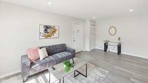 Ein modernes Wohnzimmer mit grauem Sofa, Couchtisch mit Pflanze und minimalistischer Einrichtung, darunter ein gerahmtes Kunstwerk und ein runder Spiegel.