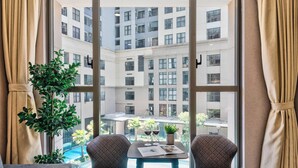 A room with a large window overlooking a courtyard with a pool and buildings. There are two chairs and a table with a plant and a menu. - S Lux Apartment (Ho Chi Minh City)