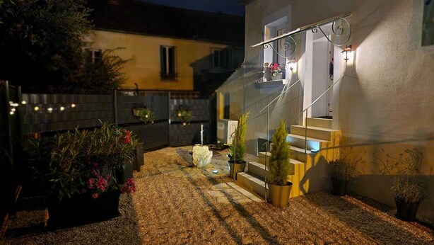 Espacio exterior nocturno con una escalera que conduce a un edificio, rodeado de plantas en macetas y un camino de grava.