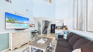 A spacious living room with a large flat-screen TV, a wooden coffee table, a brown sofa, and a kitchen in the background.