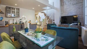 A modern living space with a glass dining table, green chairs, a blue sofa, and a kitchen area with a brick wall and a TV.