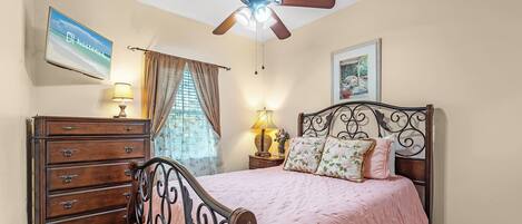 A bedroom with a bed, dresser, ceiling fan, and a TV on the wall.