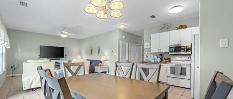 A dining area with a wooden table and chairs, a television, and a kitchen with white appliances.