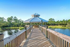A wooden boardwalk leading to a gazebo with a blue roof, surrounded by water and greenery. - Romar Lakes 104B (Orange Beach)