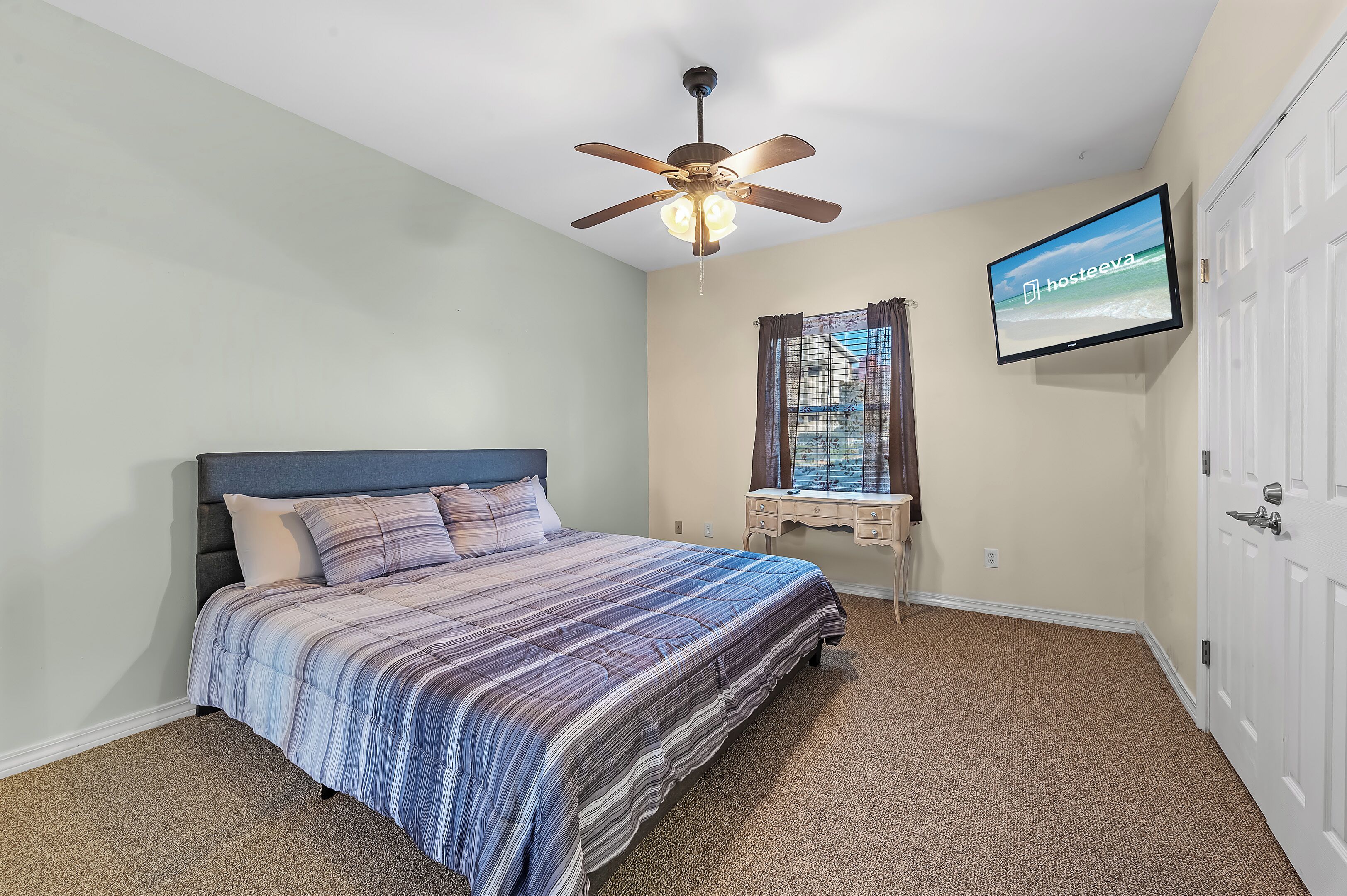 Un dormitorio con cama, ventilador de techo, televisión, ventana con cortinas y puerta.