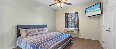 Un dormitorio con cama, ventilador de techo, televisión, ventana con cortinas y puerta.