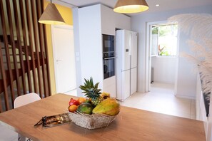 A modern kitchen with white cabinets, a refrigerator, and a dining area with a wooden table and a fruit basket. - Villa Koh-I-noor (Le Diamant)