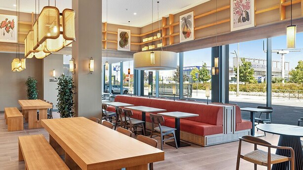 Ein Restaurant mit Holztischen und -bänken, einer roten Sitzecke mit einer Bank und großen Fenstern mit Blick auf die Straße.