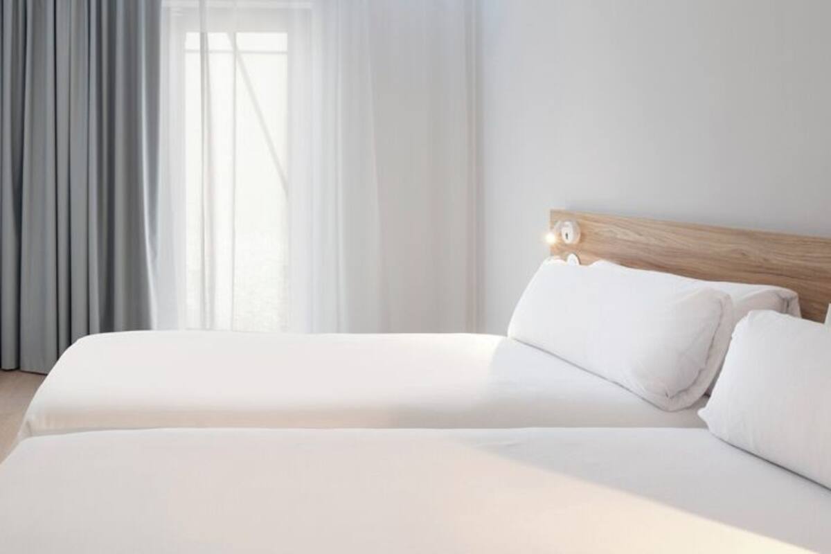 Una habitación de hotel con cama doble, cabecero de madera y un ventanal con cortinas traslúcidas.