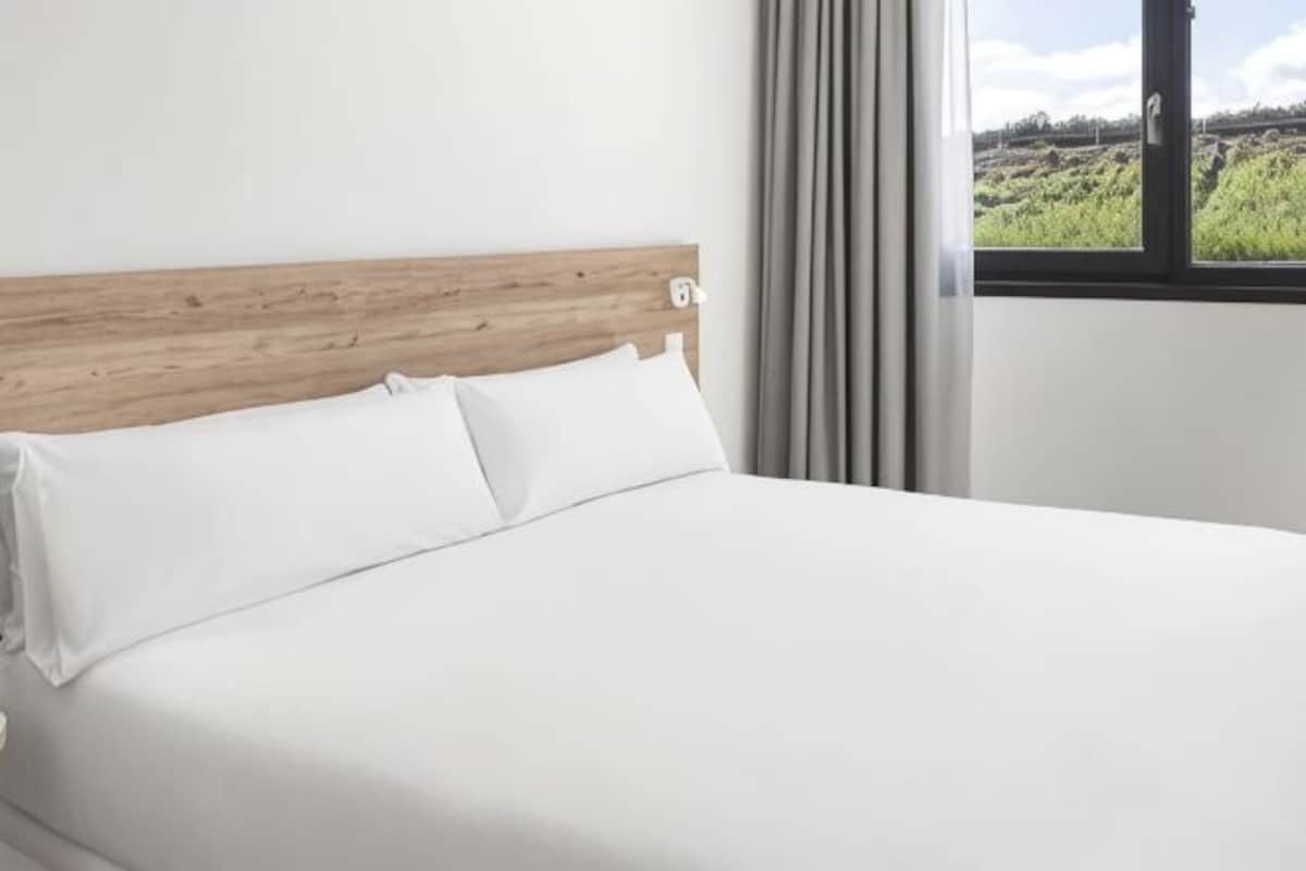 Una cama con cabecera de madera, ropa de cama blanca y ventana con vistas a un paisaje verde.