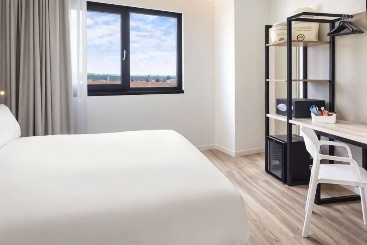 Habitación de hotel con cama, escritorio, silla, ventana con vistas y estantería.
