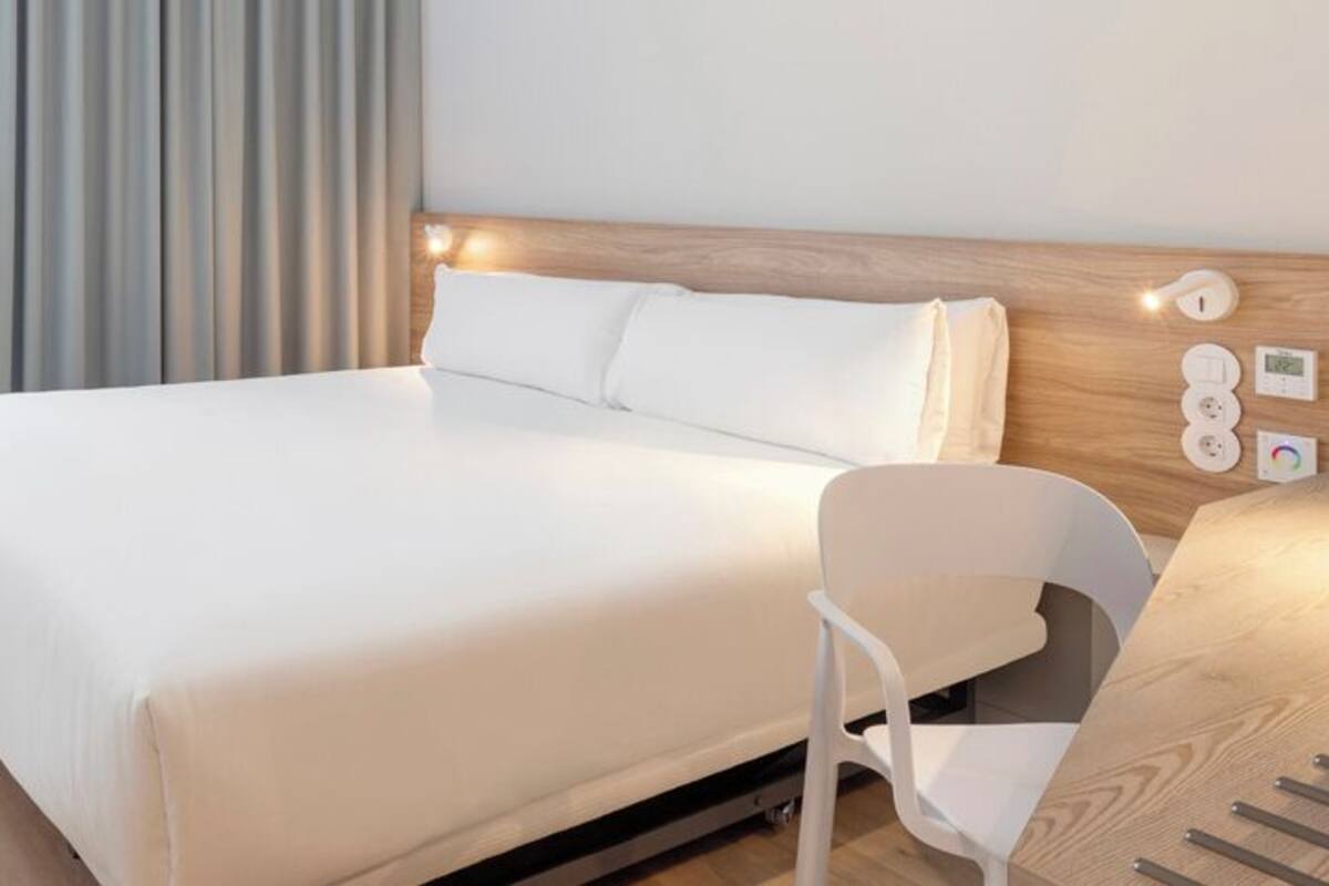 Una habitación de hotel moderna con una cama grande, una mesita de noche, una silla y un panel de control en la pared.