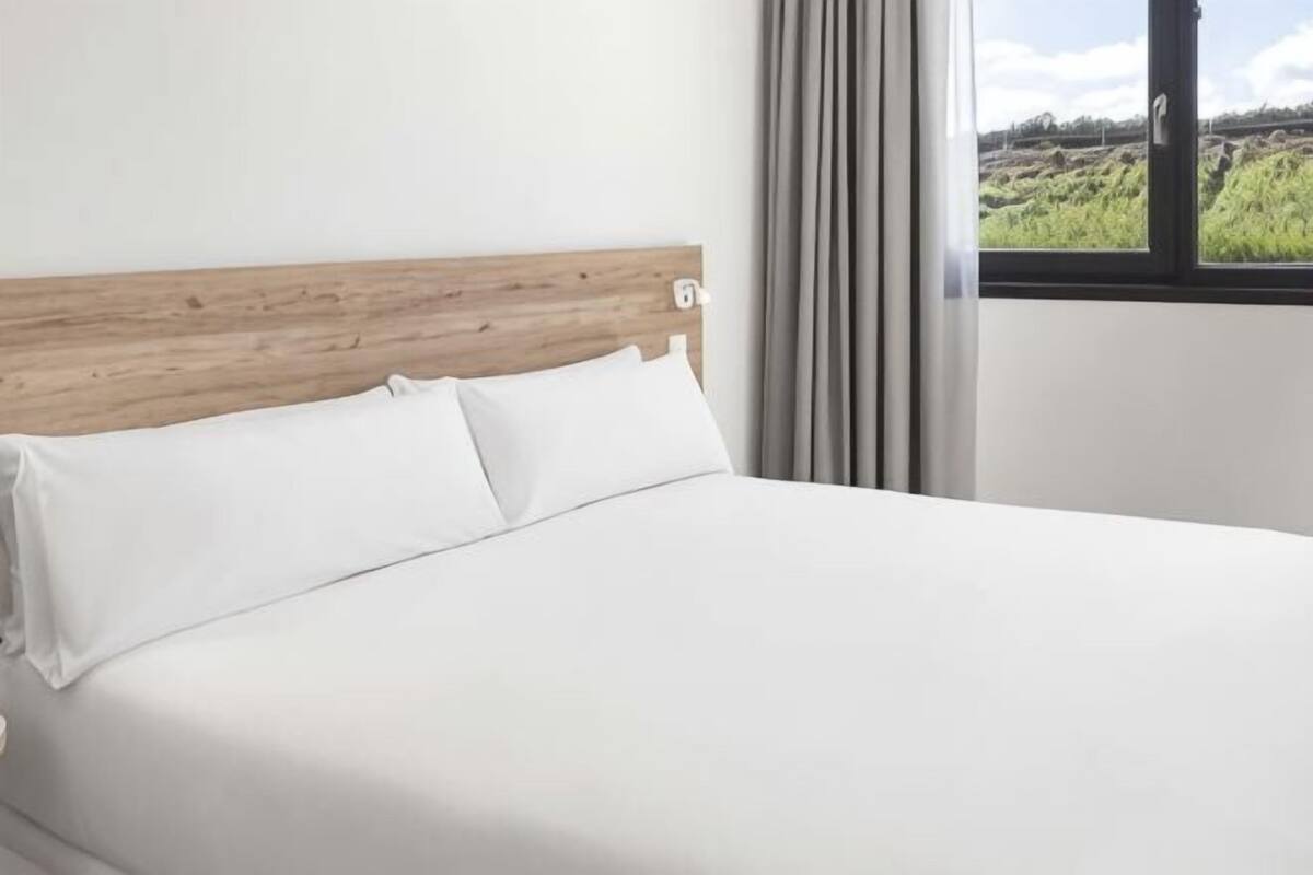 Una cama con cabecera de madera, ropa de cama blanca y ventana con vistas a un paisaje verde.