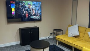 A living room with a yellow sofa, a black entertainment unit, and a TV displaying a game. - Cozy Basildon Retreat With 3 Bedrooms Free Parking (Basildon)