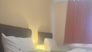 A hotel room with a bed, pillows, a nightstand with a lamp, and a window with curtains. - Cozy Basildon Retreat With 3 Bedrooms Free Parking (Basildon)